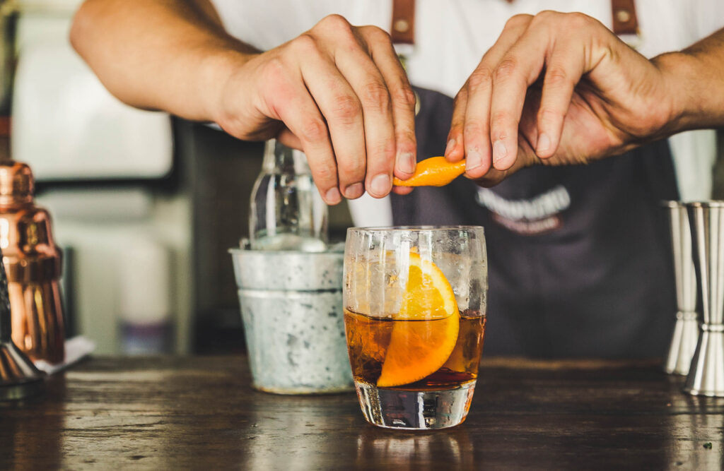 Bartender squeezes orange into a glass of iced cocktail at a bar surface in the foreground