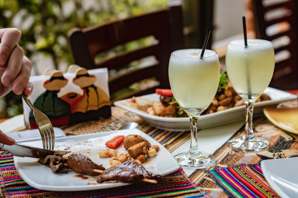 Two frosted lemonade glasses with ice and black straws on a sunny table, next to a person slicing meat at a dinning setup.
