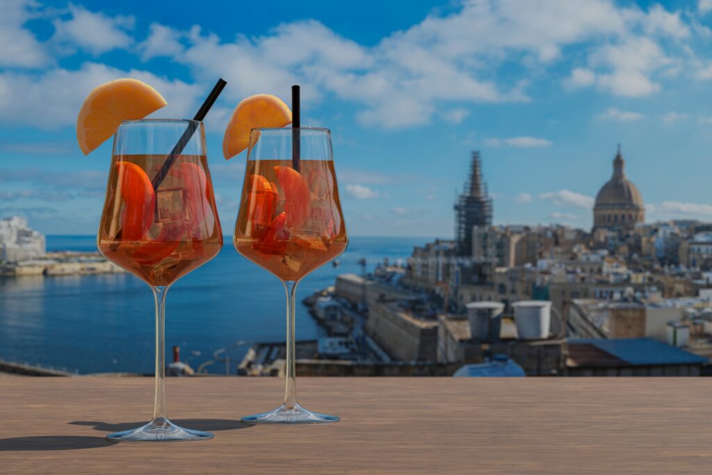 Two cocktails with orange wedges on a wooden table, overlooking a coastal city and blue sky.