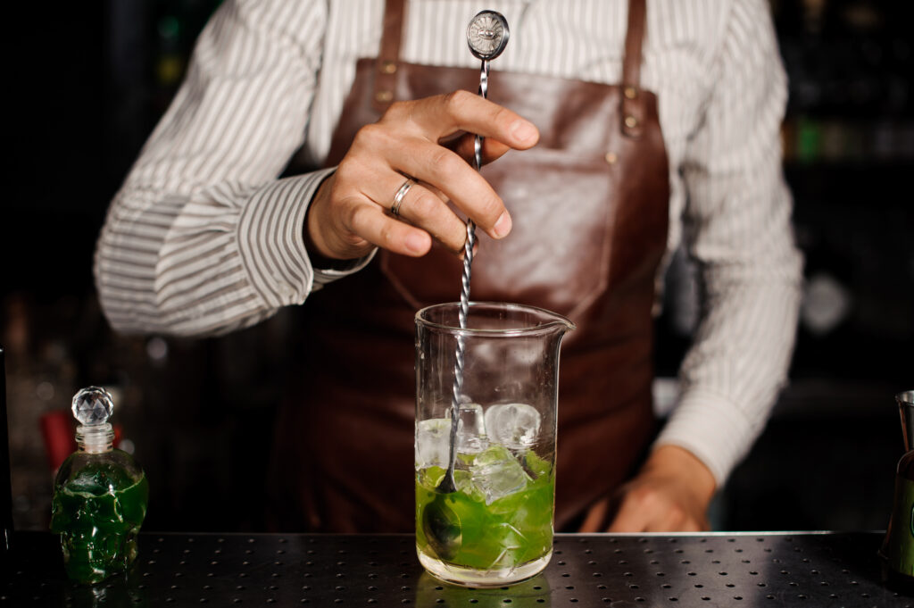 Bartender in a striped shirt and brown apron stirs a green cocktail with a long twisted spoon over a glass with ice and lime leaves.