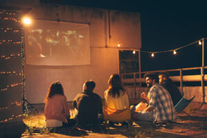 A group of friends sitting on a rooftop at night, watching a movie projected on a hanging screen, with string lights overhead