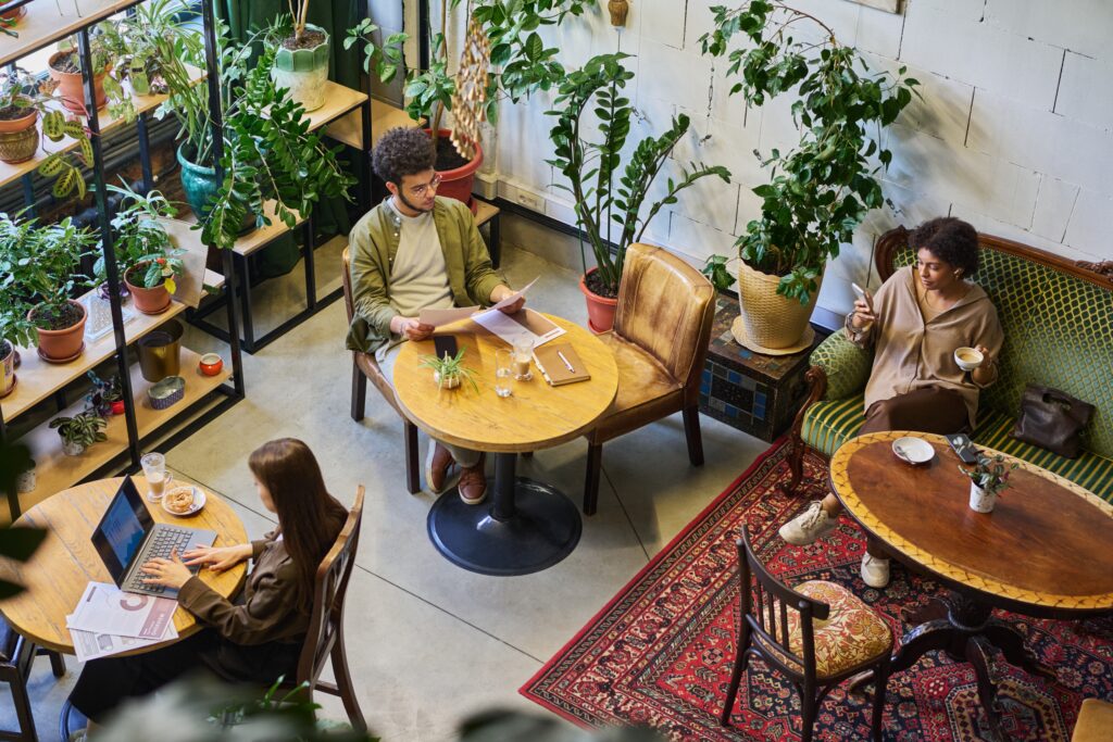 Overhead view of a plant-filled café interior with several people seated at separate wooden tables, one reading papers, one typing on a laptop, and one holding a coffee cup while looking at their phone.