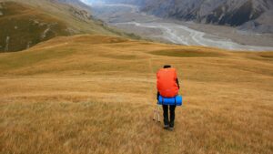 Hiker with a large orange backpack walking across golden grassland toward a wide mountain valley with a braided river