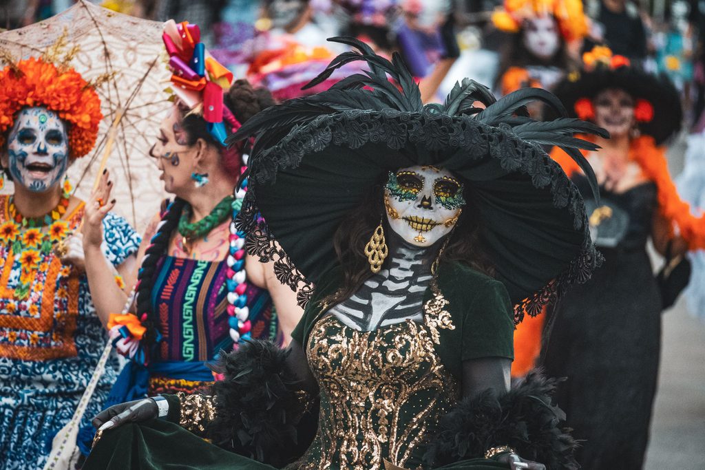 pexels-dany-arriaga-629175657-34702975 - Couchsurfing Blog Alt text: A woman in skeleton face paint, a wide black feathered hat, and a green and gold gown walking in a parade, with other costumed marchers behind her