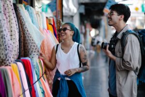A woman with blue hair and sunglasses laughing as she touches colorful fabrics at a market stall, while a man with a camera and backpack smiles beside her.