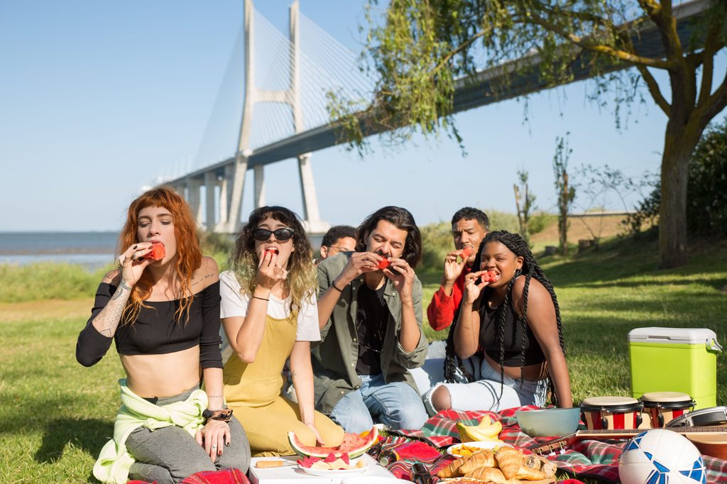 A group of friends sharing watermelon on a picnic blanket in a park by the water, with a large bridge behind them