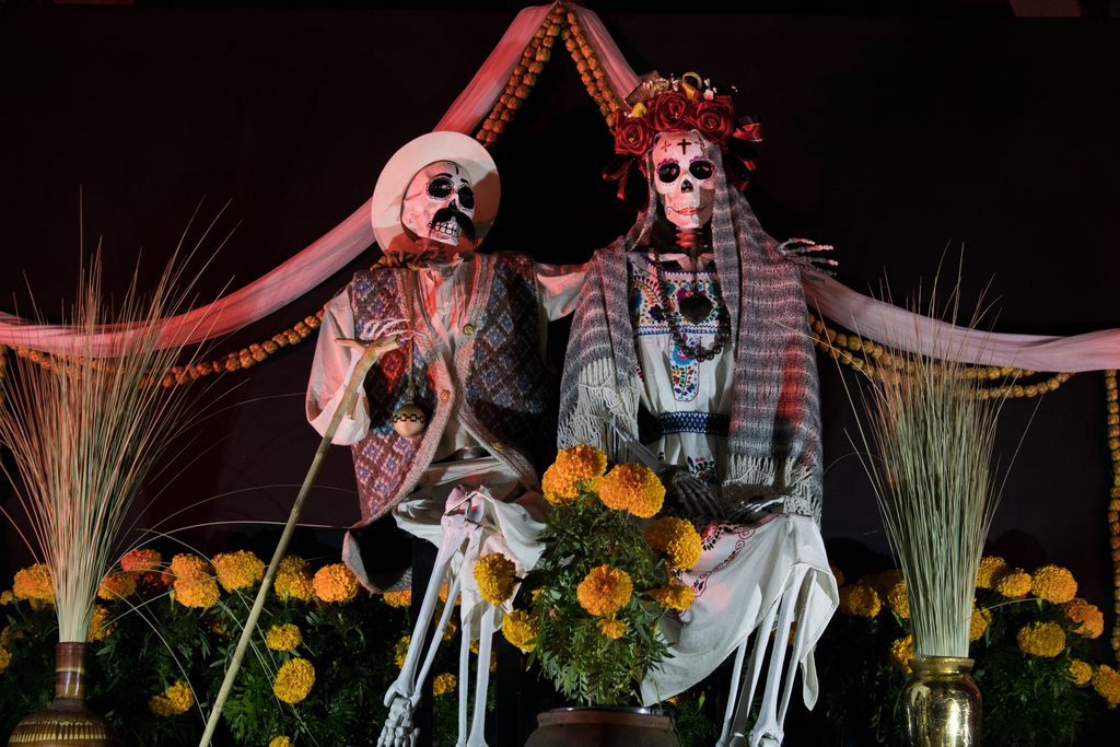 pexels-marco-ramos-223569157-14302939 - Couchsurfing Blog Two skeleton figures in traditional clothing seated among bright orange marigolds under red draped fabric on an altar display