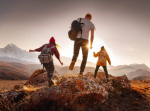 Three friends with backpacks stepping across lichen-covered rocks toward a setting sun, with snow-capped mountains in the distance.