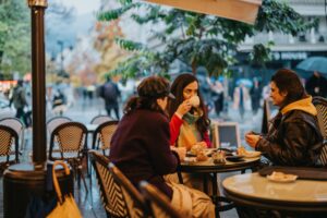 Three women sitting at an outdoor café table, talking and drinking coffee on a rainy city street.