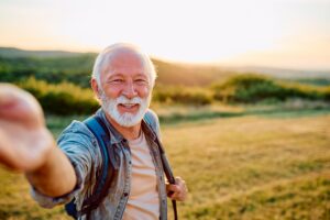 Smiling older man with white hair and a beard, wearing a denim shirt and blue backpack, reaching toward the camera for a selfie in a grassy field at sunset